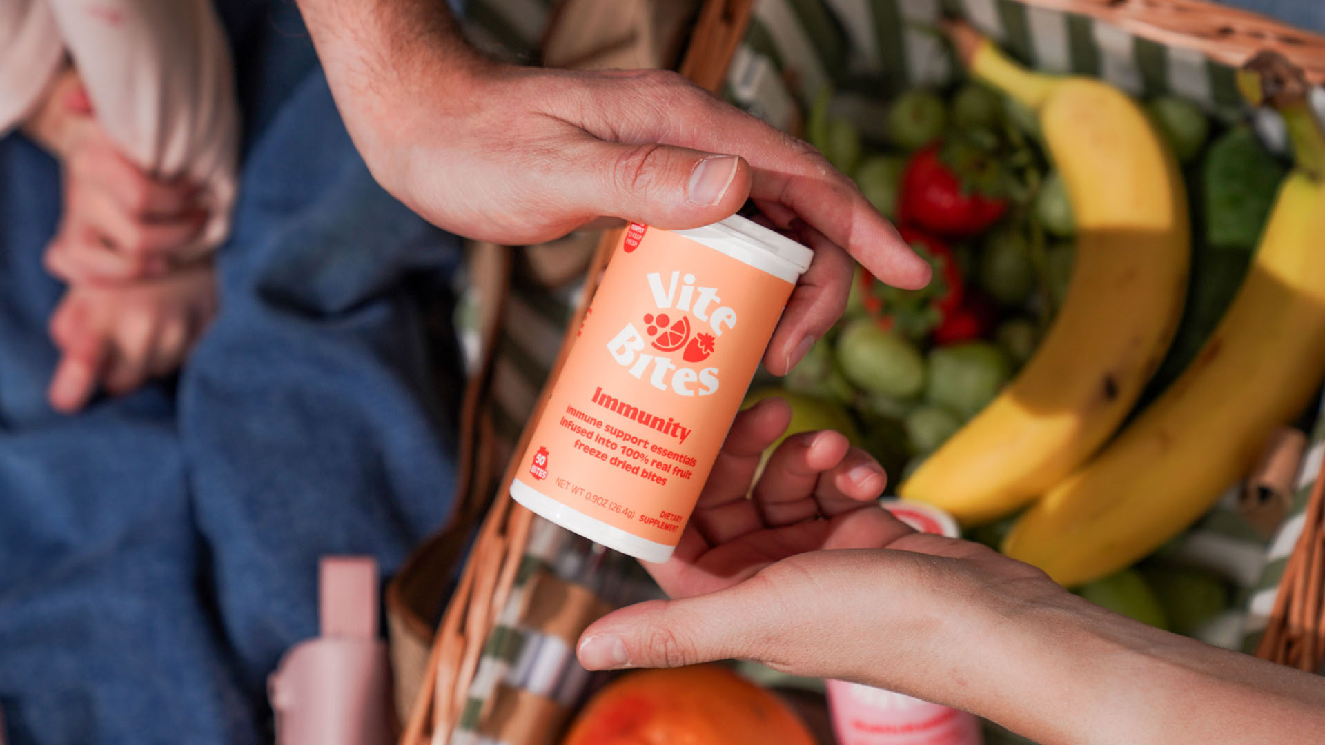 ViteBites Immunity Lifestyle Hands holding a ViteBites Immunity supplement container above a basket of fresh fruit